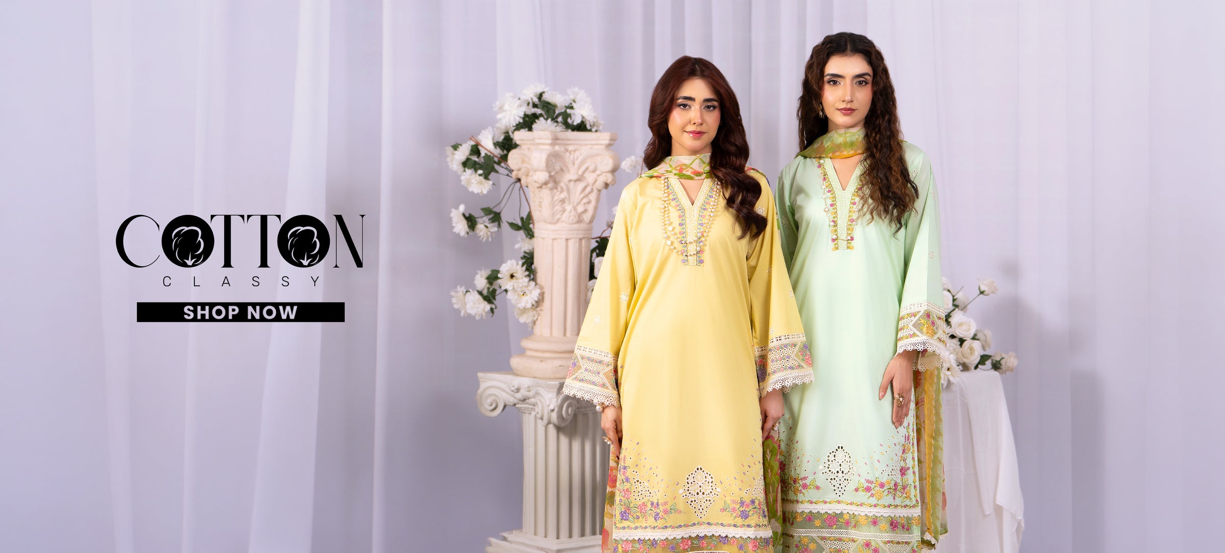Two women wearing light green and yellow embroidered dresses with floral decorations in the background.